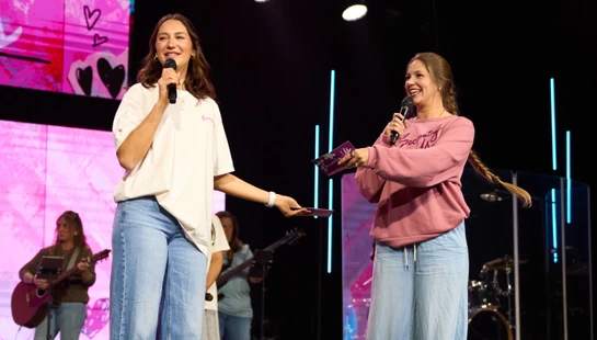 Two women stand on a stage holding microphones and smiling. In the background, a band is performing in front of a large pink screen with heart graphics.